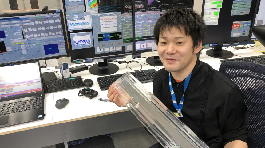 Ichiro Inoue working in the control room of SACLA (Japanese X-ray free-electron laser facility), where he is controlling the machine to generate twin XFEL pulses. He is holding an X-ray mirror for focusing the X-ray beam to micrometer size. (Credit: SACLA / IFJ PAN)